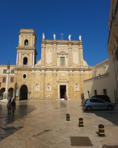 A large, light-colored stone cathedral with a single bell tower on the left, a central facade featuring statues along the roofline and niches with statues below, situated in a paved public square under a clear blue sky.