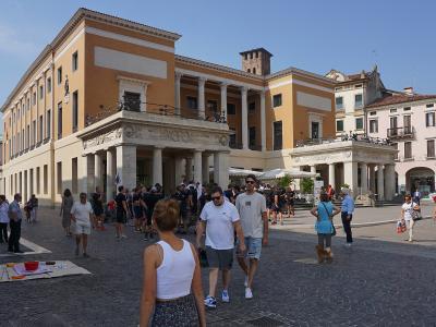 A large neoclassical building with a colonnaded portico and yellow facade, situated in a bustling cobblestone piazza with many pedestrians and outdoor seating.