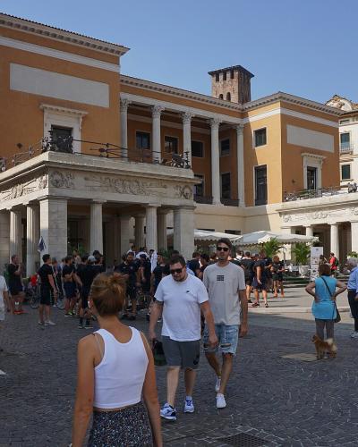 A large neoclassical building with a colonnaded portico and yellow facade, situated in a bustling cobblestone piazza with many pedestrians and outdoor seating.