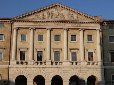 The image shows the neoclassical facade of a theater with six large columns, a pediment featuring sculptural reliefs, and the inscription "TEATRO DELLE MVSE" carved into the entablature; the building is constructed of light-colored stone or stucco