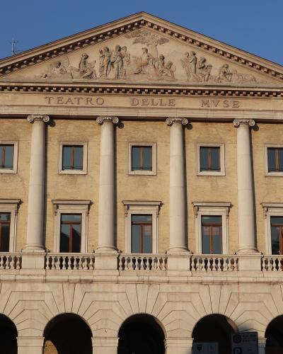 The image shows the neoclassical facade of a theater with six large columns, a pediment featuring sculptural reliefs, and the inscription "TEATRO DELLE MVSE" carved into the entablature; the building is constructed of light-colored stone or stucco