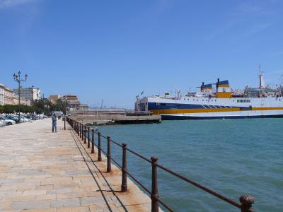 A wide-angle view of a paved waterfront promenade lined with parked cars and ornate streetlamps, with a large white and blue ferry docked at a pier in turquoise water under a clear blue sky.