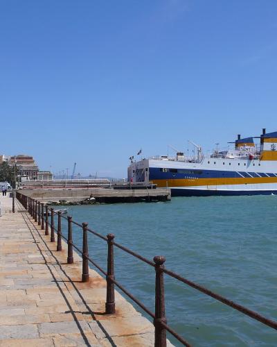 A wide-angle view of a paved waterfront promenade lined with parked cars and ornate streetlamps, with a large white and blue ferry docked at a pier in turquoise water under a clear blue sky.