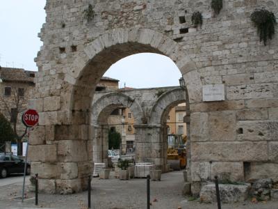 A large, ancient stone gate with a prominent central arch and two smaller side arches, featuring a white plaque on the right pillar that reads "CARRO PORTA ROMANA", set against a street scene with a stop sign and construction equipment.