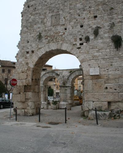 A large, ancient stone gate with a prominent central arch and two smaller side arches, featuring a white plaque on the right pillar that reads "CARRO PORTA ROMANA", set against a street scene with a stop sign and construction equipment.