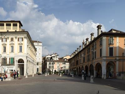 A wide-angle view of a sunlit Italian piazza featuring a large yellow building with arched porticoes on the left, a statue on a pedestal, and a long orange building with a row of chimneys and arches on the right, under a partly cloudy sky.