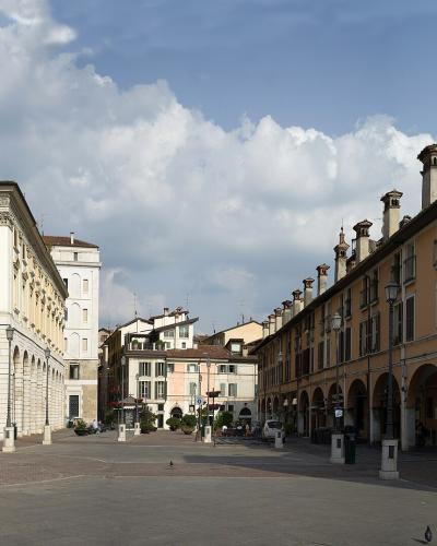 A wide-angle view of a sunlit Italian piazza featuring a large yellow building with arched porticoes on the left, a statue on a pedestal, and a long orange building with a row of chimneys and arches on the right, under a partly cloudy sky.
