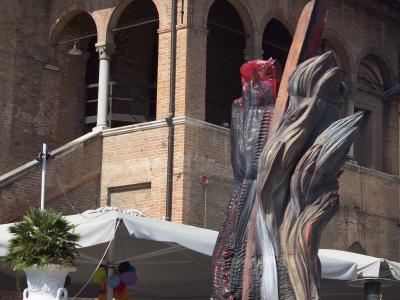 A large, abstract, flame-like sculpture with red and metallic tones stands on a stone pedestal in front of a historic brick building with arched loggias, near an outdoor cafe with white umbrellas.