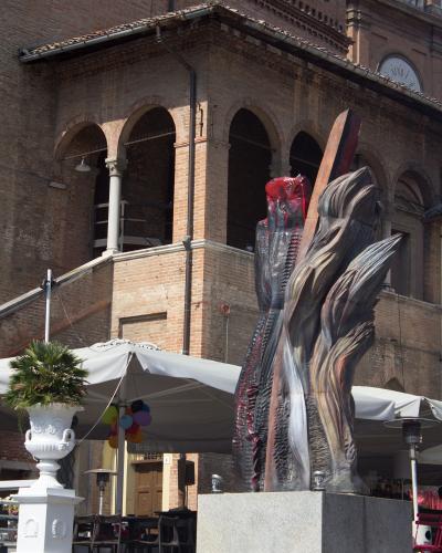 A large, abstract, flame-like sculpture with red and metallic tones stands on a stone pedestal in front of a historic brick building with arched loggias, near an outdoor cafe with white umbrellas.