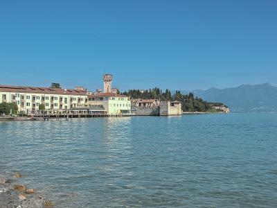 A view across a lake of a peninsula featuring a prominent medieval stone tower and fortified walls adjacent to modern buildings, with mountains in the background.