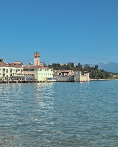 A view across a lake of a peninsula featuring a prominent medieval stone tower and fortified walls adjacent to modern buildings, with mountains in the background.