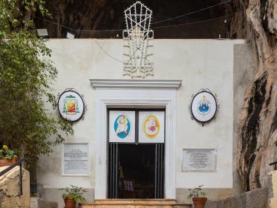 The image shows the entrance to a small white-walled chapel built into the base of a rocky cliff face, featuring religious plaques and a metal cross structure above the door.