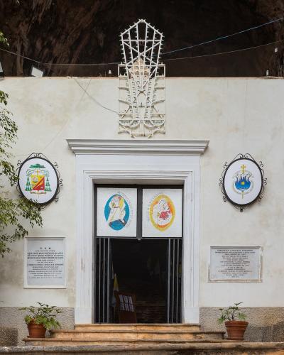 The image shows the entrance to a small white-walled chapel built into the base of a rocky cliff face, featuring religious plaques and a metal cross structure above the door.