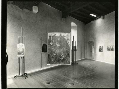 A black-and-white photograph of an interior gallery space featuring medieval-style paintings displayed on modern metal easels and wall mounts, with rough plaster walls, wooden floors, and exposed dark ceiling beams.
