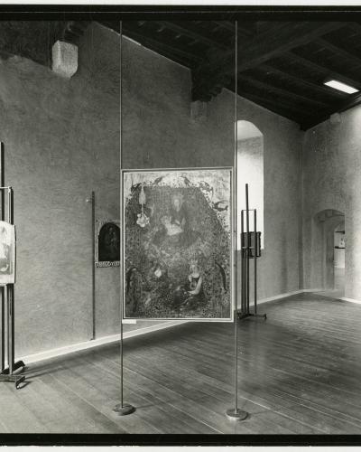 A black-and-white photograph of an interior gallery space featuring medieval-style paintings displayed on modern metal easels and wall mounts, with rough plaster walls, wooden floors, and exposed dark ceiling beams.