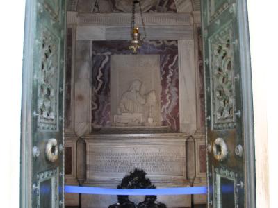 The image shows the interior of a marble tomb monument viewed through open green doors, featuring a relief sculpture of a seated figure reading, an inscription plaque below it, and a wreath at the base.