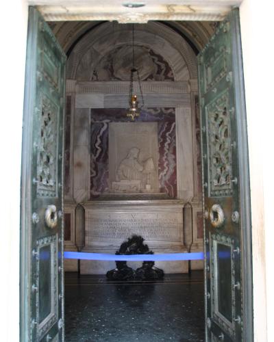 The image shows the interior of a marble tomb monument viewed through open green doors, featuring a relief sculpture of a seated figure reading, an inscription plaque below it, and a wreath at the base.