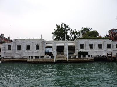 The image shows the exterior of a white neoclassical building situated directly on a canal, with signage reading "PEGGY GUGGENHEIM COLLECTION" and a poster for a "PICASSO" exhibition; two sphinx statues flank the entrance steps leading down to the