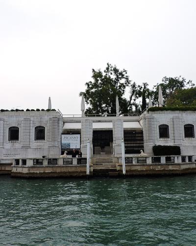 The image shows the exterior of a white neoclassical building situated directly on a canal, with signage reading "PEGGY GUGGENHEIM COLLECTION" and a poster for a "PICASSO" exhibition; two sphinx statues flank the entrance steps leading down to the