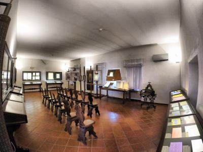 The image shows the interior of a museum exhibit featuring rows of antique wooden chairs, display cases with papers or documents, and historical papermaking equipment like a hand-cranked machine, with signage visible in the background.