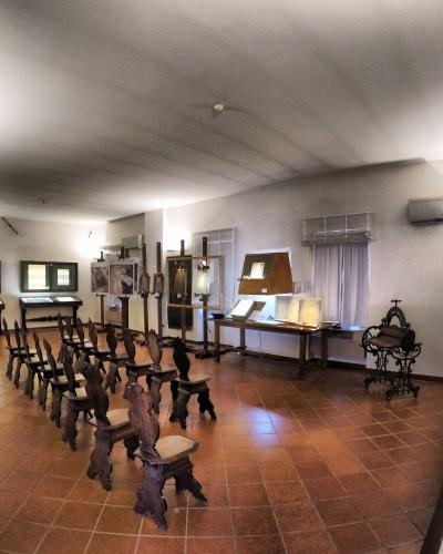The image shows the interior of a museum exhibit featuring rows of antique wooden chairs, display cases with papers or documents, and historical papermaking equipment like a hand-cranked machine, with signage visible in the background.