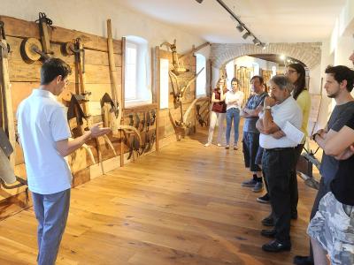 A group of people are gathered in a museum gallery with wooden floors and walls, listening to a guide who is gesturing toward a display of traditional wooden and metal agricultural tools mounted on the wall.