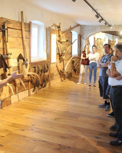A group of people are gathered in a museum gallery with wooden floors and walls, listening to a guide who is gesturing toward a display of traditional wooden and metal agricultural tools mounted on the wall.