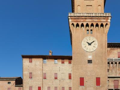 A tall brick tower with a large clock face, crenellated battlements, and red-shuttered windows, part of a larger medieval brick fortress complex under a clear blue sky.