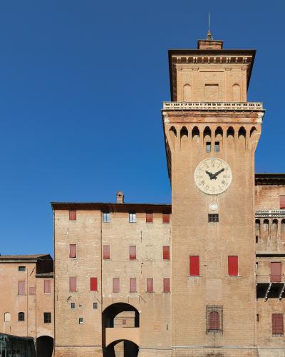 A tall brick tower with a large clock face, crenellated battlements, and red-shuttered windows, part of a larger medieval brick fortress complex under a clear blue sky.