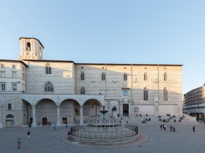 The image shows a large, sunlit public square featuring a prominent medieval stone building with Gothic windows and a loggia, centered around a circular fountain, with people scattered across the paved area.
