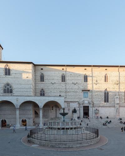 The image shows a large, sunlit public square featuring a prominent medieval stone building with Gothic windows and a loggia, centered around a circular fountain, with people scattered across the paved area.