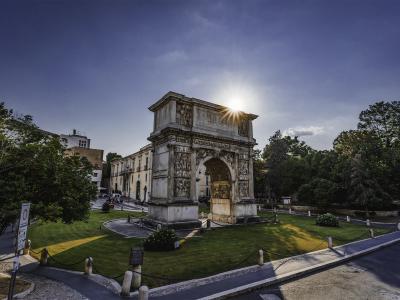 A large, single-arched Roman triumphal arch with extensive relief sculptures on its piers and attic, situated in a grassy park-like setting with trees and a road in the foreground, under a bright sunburst.