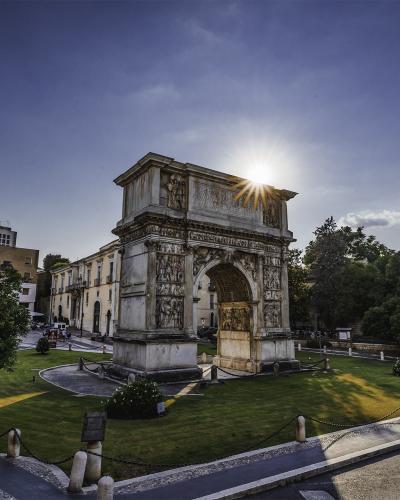A large, single-arched Roman triumphal arch with extensive relief sculptures on its piers and attic, situated in a grassy park-like setting with trees and a road in the foreground, under a bright sunburst.