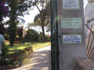 A sunlit garden entrance with a stone pillar displaying three signs: the top one reads "GIARDINI AUGUSTO," the middle one is from the "MUNICIPIO di CAPRI" prohibiting flower picking, and the bottom one promotes cleanliness and silence; a brick pat