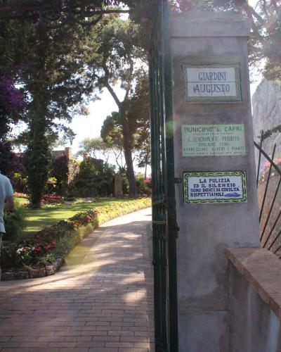 A sunlit garden entrance with a stone pillar displaying three signs: the top one reads "GIARDINI AUGUSTO," the middle one is from the "MUNICIPIO di CAPRI" prohibiting flower picking, and the bottom one promotes cleanliness and silence; a brick pat