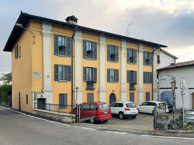 A large, yellow, multi-story building with white pilasters and blue shutters, situated on a street corner with parked cars and a sign reading "CORTILE PALASIO" on the facade.