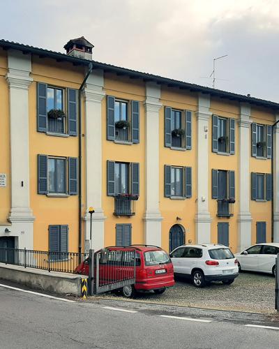 A large, yellow, multi-story building with white pilasters and blue shutters, situated on a street corner with parked cars and a sign reading "CORTILE PALASIO" on the facade.