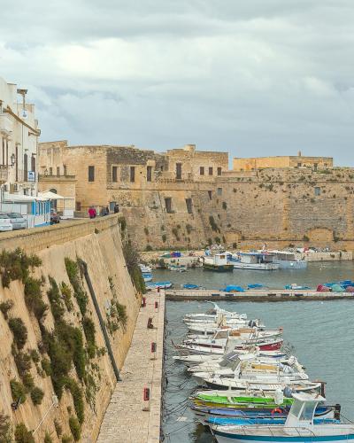 The image shows a coastal harbor scene with a row of parked cars along a stone seawall, a line of small fishing boats moored in the water, and a large, historic stone fortress with rounded bastions in the background under an overcast sky.