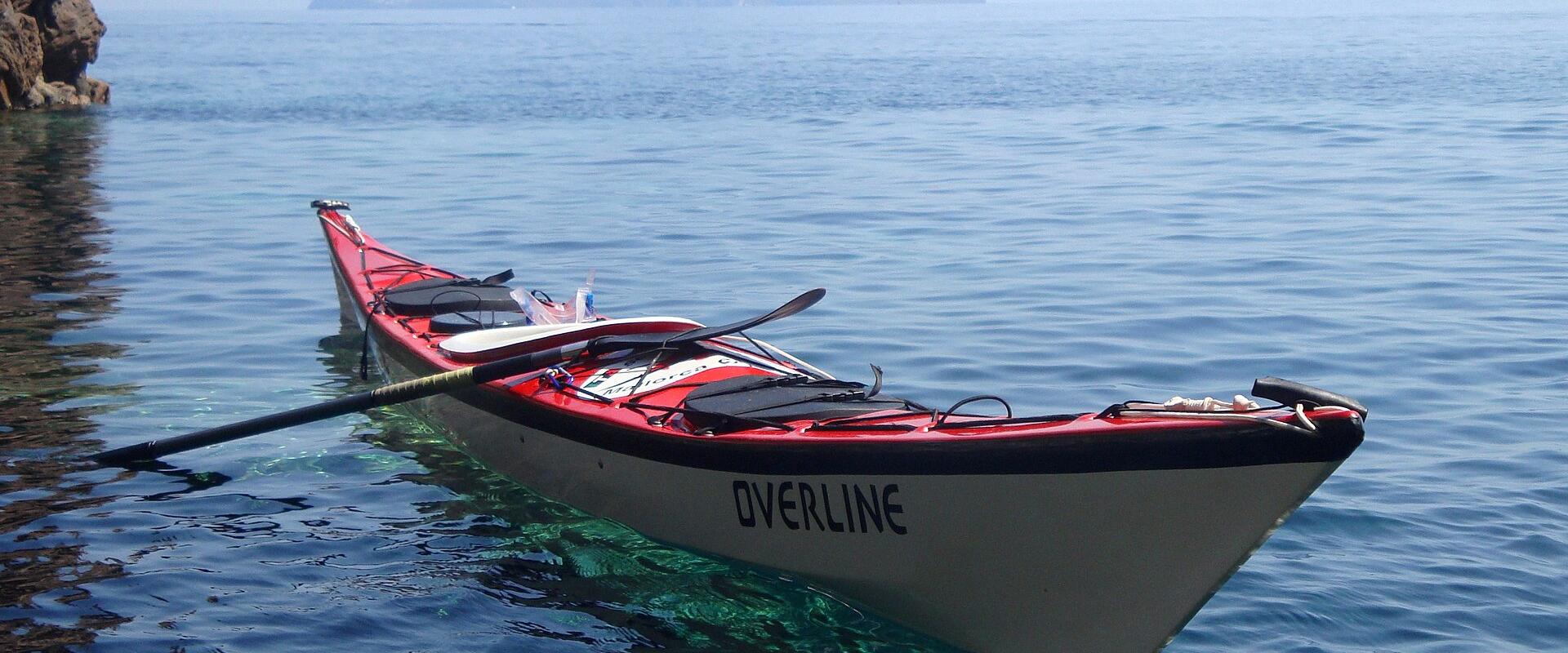 A red and white sea kayak with "OVERLINE" written on its side floats in calm blue water near a rocky cliff, with a large conical volcanic island visible in the background under a clear sky.
