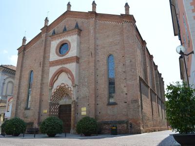 A low-angle exterior shot of a large, red-brick Gothic-style church with a prominent arched entrance featuring checkered stonework, a circular rose window above the portal, and multiple tall narrow windows; the building is situated on a paved plaz