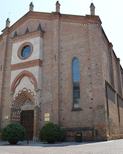 A low-angle exterior shot of a large, red-brick Gothic-style church with a prominent arched entrance featuring checkered stonework, a circular rose window above the portal, and multiple tall narrow windows; the building is situated on a paved plaz