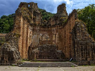 The image shows the roofless stone ruins of a historic church or monastery with a prominent cross on the apse wall, set against a forested hillside.