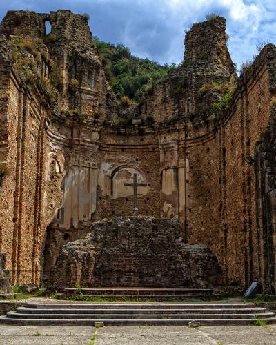 The image shows the roofless stone ruins of a historic church or monastery with a prominent cross on the apse wall, set against a forested hillside.