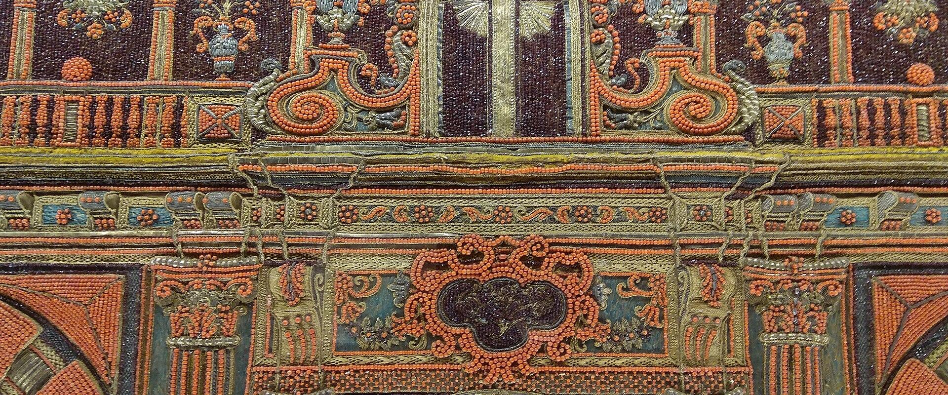 A close-up photograph of an ornate, beaded textile or embroidery featuring architectural motifs like arches, columns, and a central cross, with visible textural detail of individual beads.
