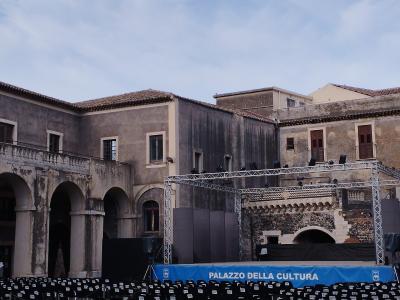 A courtyard of an old stone building with arched colonnades, featuring a temporary stage setup with lighting trusses and rows of black chairs, with a blue banner reading "PALAZZO DELLA CULTURA" in the foreground.