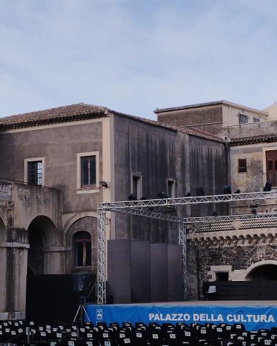 A courtyard of an old stone building with arched colonnades, featuring a temporary stage setup with lighting trusses and rows of black chairs, with a blue banner reading "PALAZZO DELLA CULTURA" in the foreground.