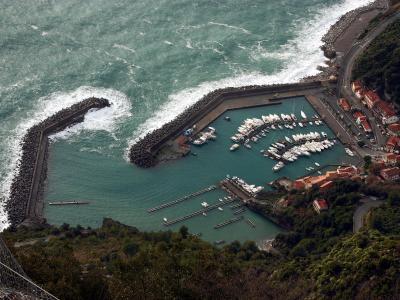 A high-angle view of a coastal harbor with a breakwater, numerous white boats docked in turquoise water, and a hillside town with red-roofed buildings; the foreground shows green vegetation and part of a wire mesh structure.