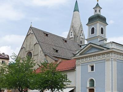 The image shows a large, light-blue Baroque-style church with a prominent tower and spire, situated in a cobblestone plaza with people and trees; behind it rises a taller, older Gothic-style church with a distinctive steep, grey-tiled roof and a p
