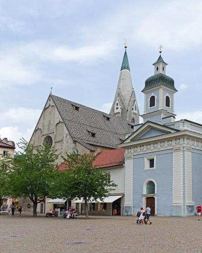 The image shows a large, light-blue Baroque-style church with a prominent tower and spire, situated in a cobblestone plaza with people and trees; behind it rises a taller, older Gothic-style church with a distinctive steep, grey-tiled roof and a p