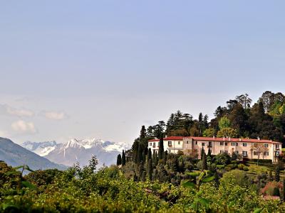 A long, pale villa with a red-tiled roof sits on a lush, tree-covered hillside, with snow-capped mountains visible in the distance across a body of water.